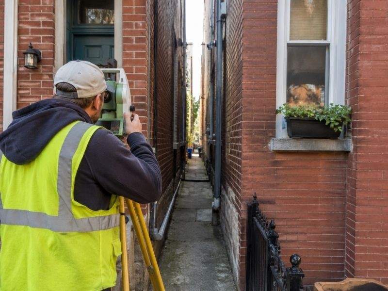 Surveyor measuring a narrow alley between attached brick rowhouses to verify property boundaries and shared access rights.